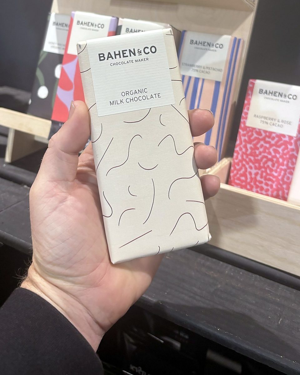 A hand holding a Bahen & Co organic milk chocolate bar in front of a display of Bahen & Co bars at Origins Market, Busselton.