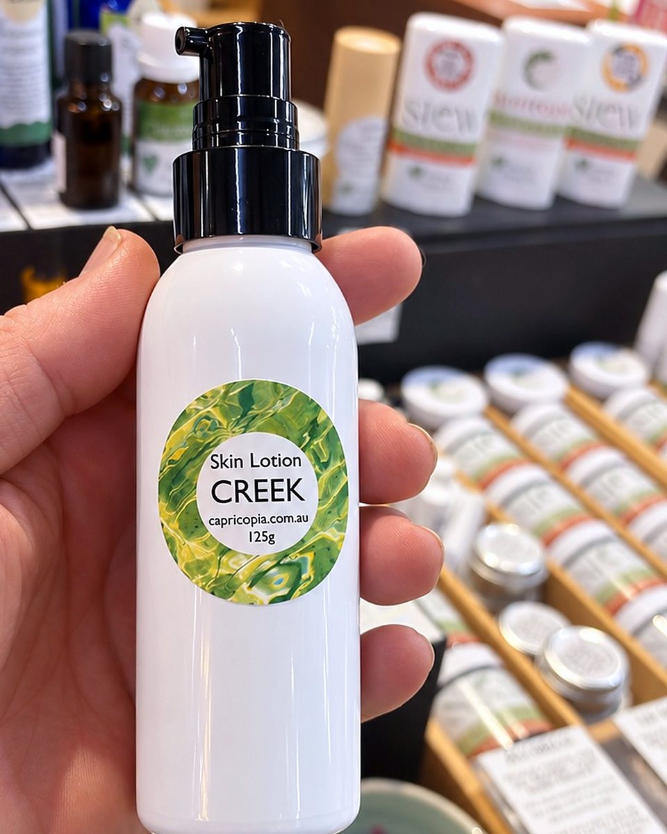 A hand holding a white pump bottle of Capricopia Creek skin lotion, with a green label, in front of a skincare display at Origins Market, Busselton.