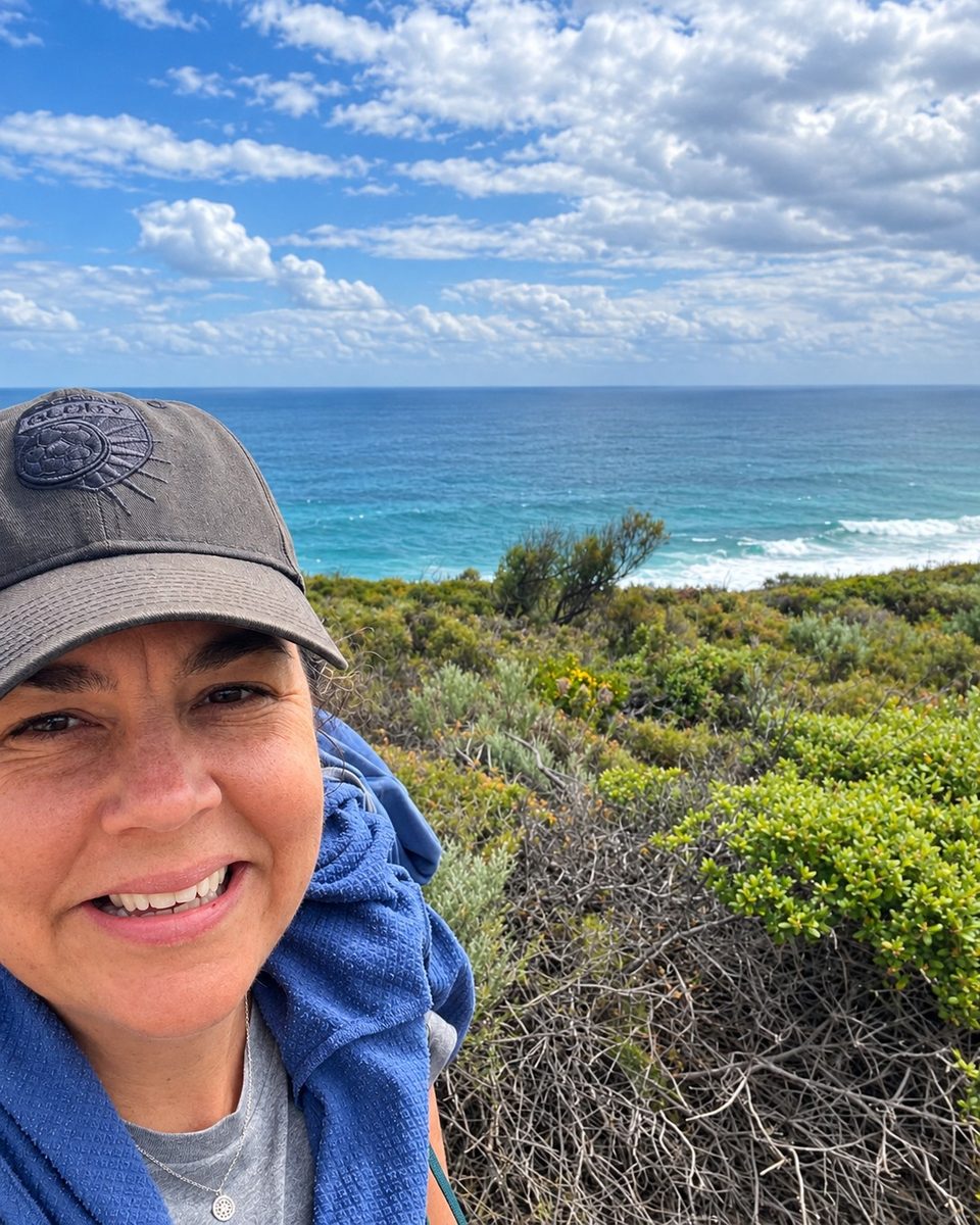 Frances smiling on the Cape to Cape Track, with coastal heath and the Indian Ocean behind her.
