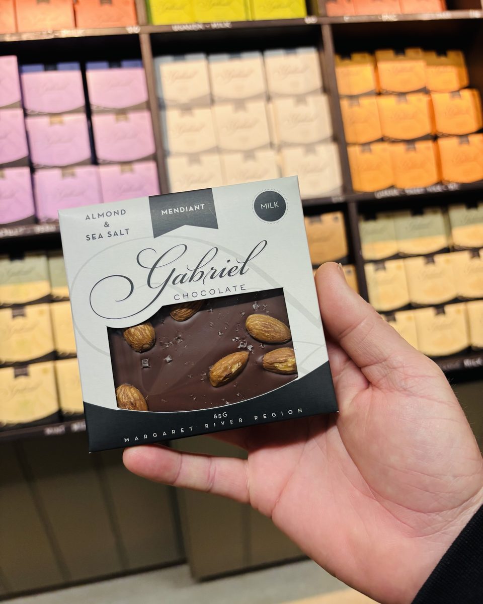 A hand holding a Gabriel Chocolate almond and sea salt bar inside the Gabriel Chocolate shop, coloured boxes on the shelves behind.