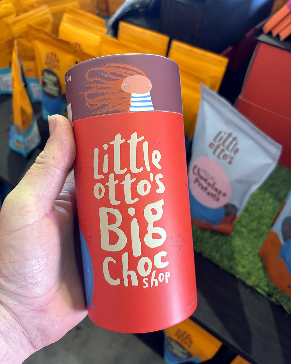 A hand holding a red Little Otto's Big Choc Shop gift tube, with the maker's chocolate bags on shelves behind, in the shop in Busselton.