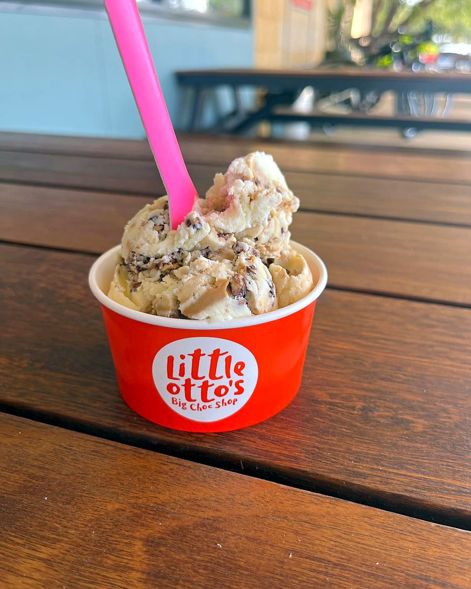A cup of gelato with a pink spoon in a red Little Otto's Big Choc Shop cup, on a timber table outside the shop in Busselton.